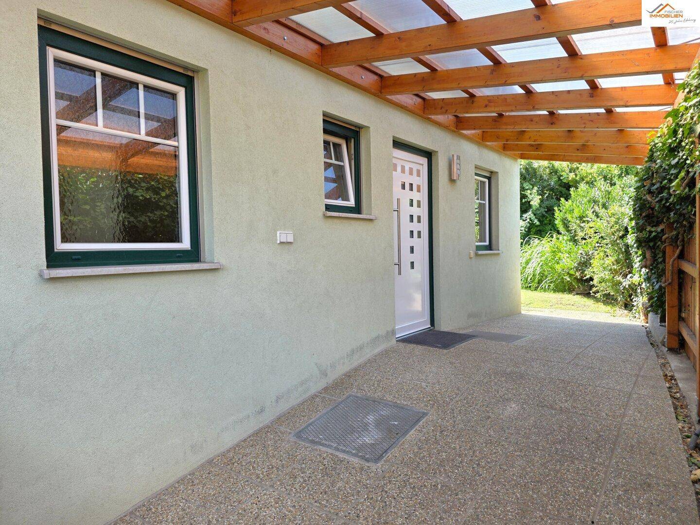 Überdachter Eingangsbereich des Hauses mit gepflastertem Weg und grünen Fensterrahmen.