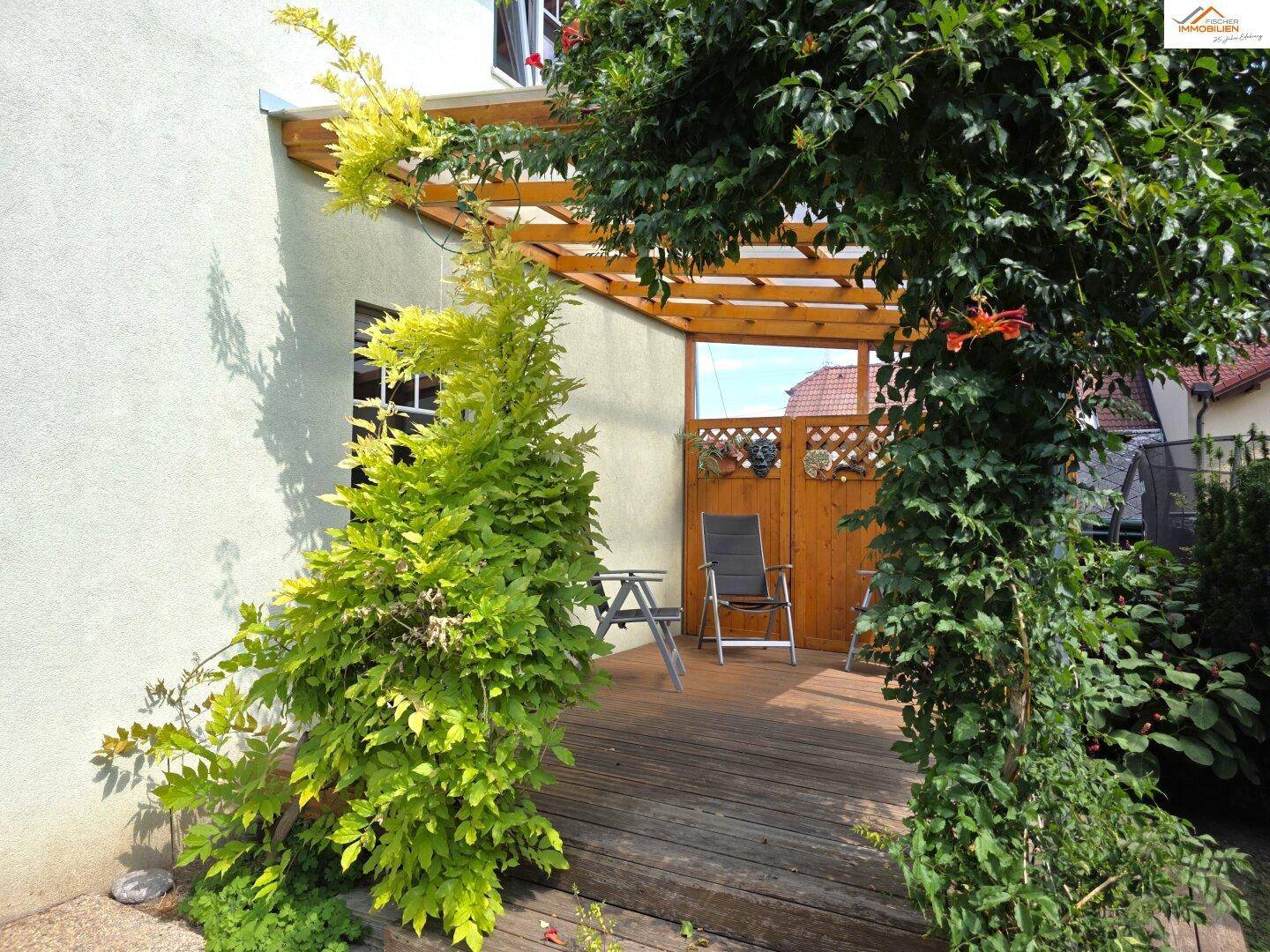Gemütliche überdachte Holzterrasse mit Sitzgelegenheiten und grüner Bepflanzung.