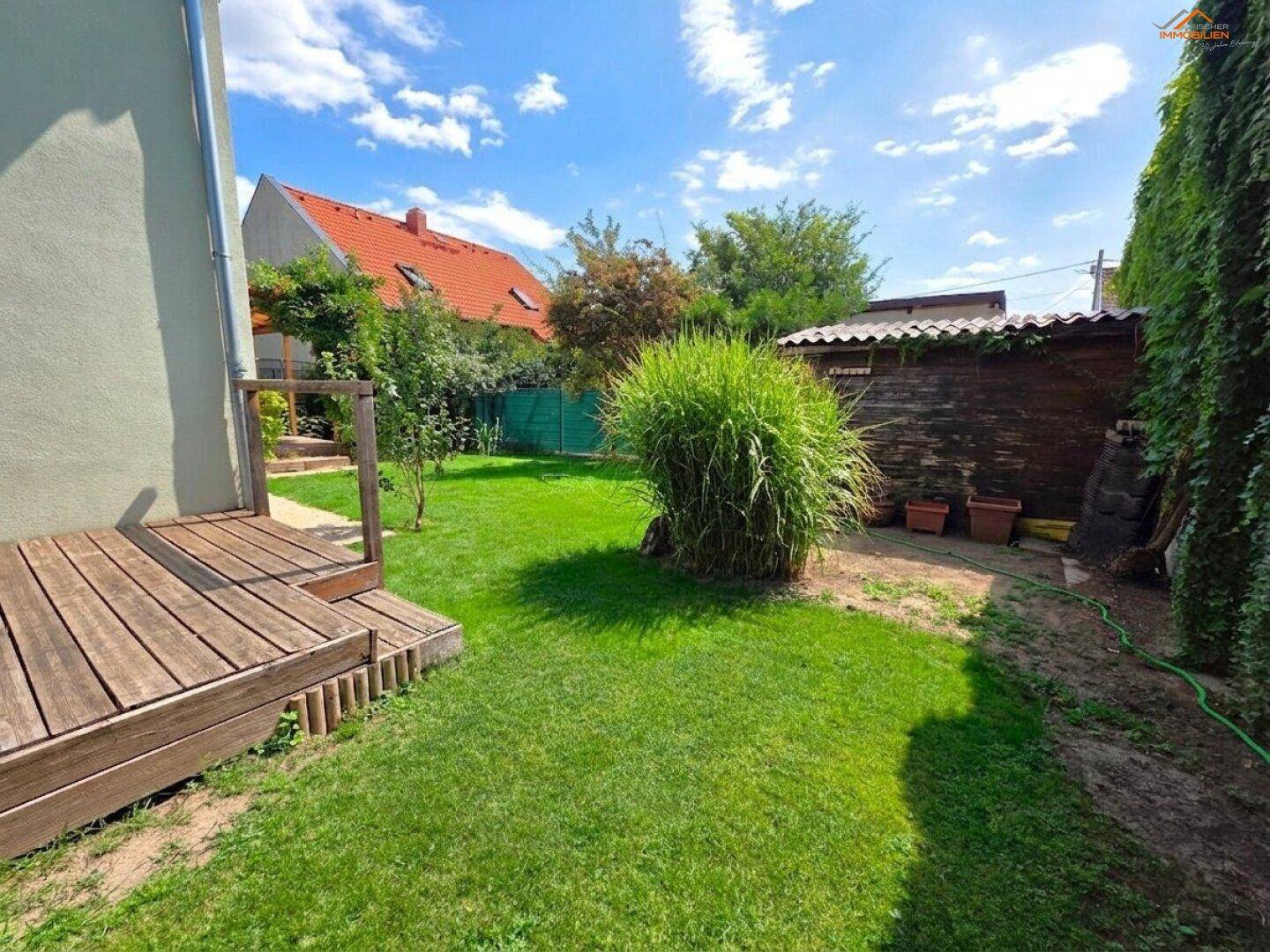 Sonniger Gartenbereich mit Rasenfläche, Holzterrasse und einem kleinen Geräteschuppen.