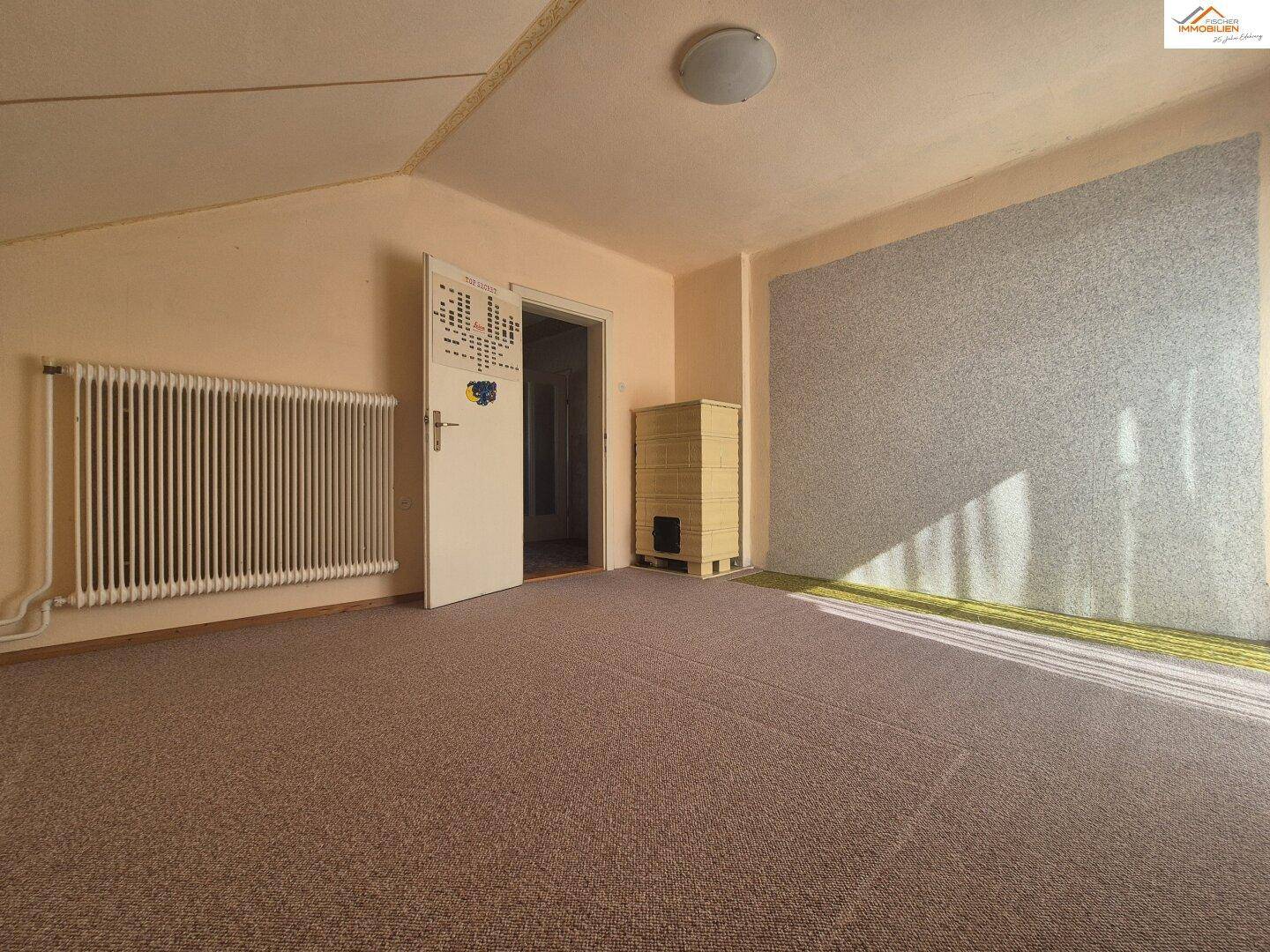 Geräumiges Zimmer mit Teppichboden, Heizkörper und einem traditionellen Kachelofen.