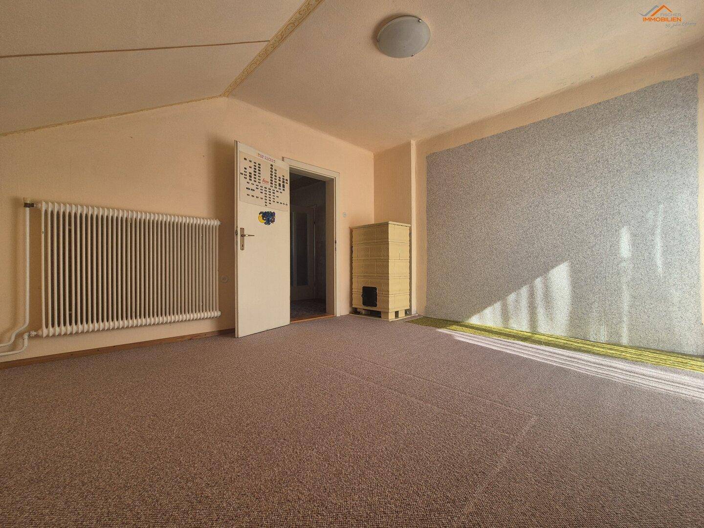 Geräumiges Zimmer mit Teppichboden, Heizkörper und einem traditionellen Kachelofen.