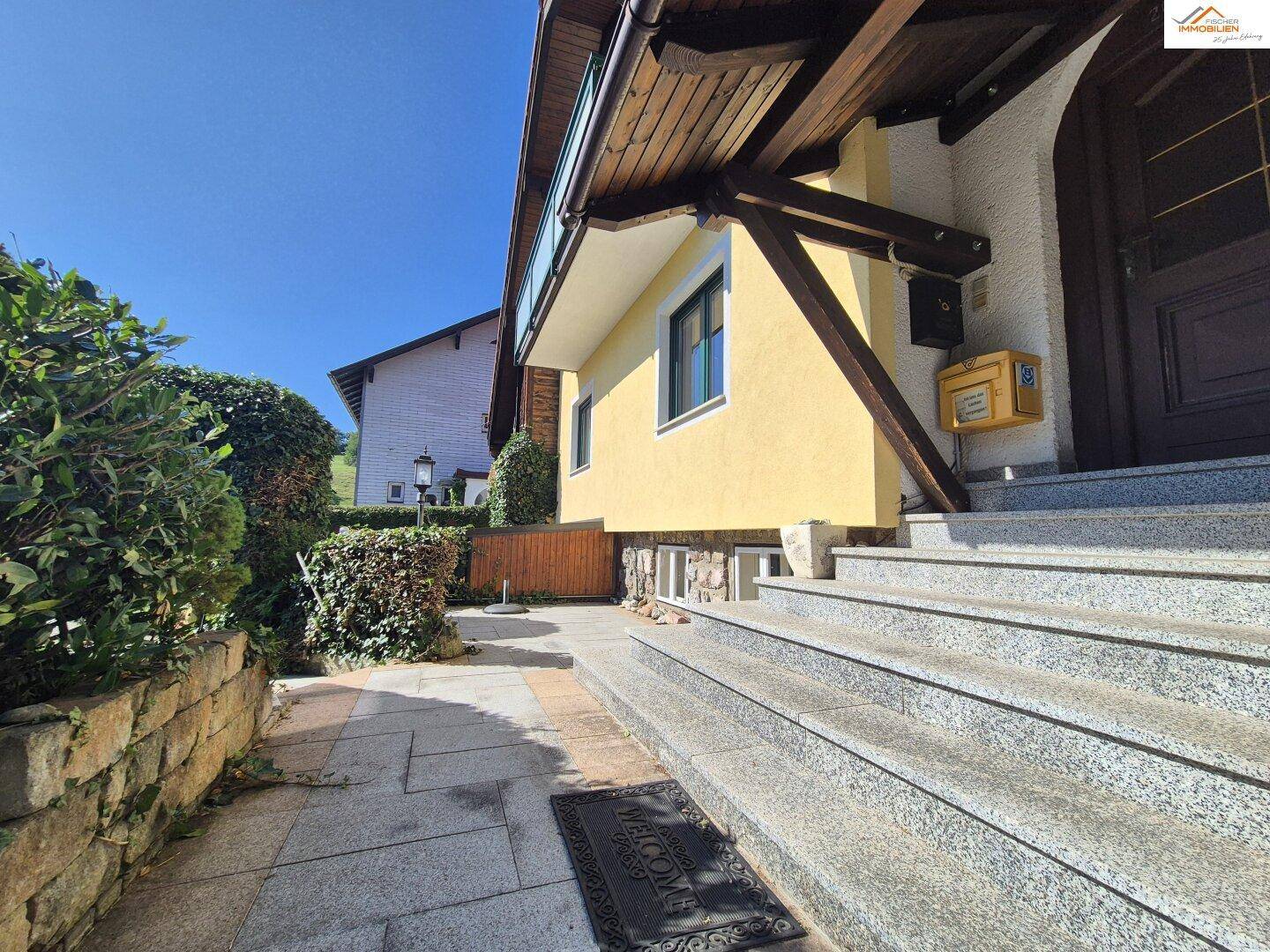 Seitlicher Eingangsbereich des Hauses mit Steintreppe, gepflastertem Weg und grüner Hecke.
