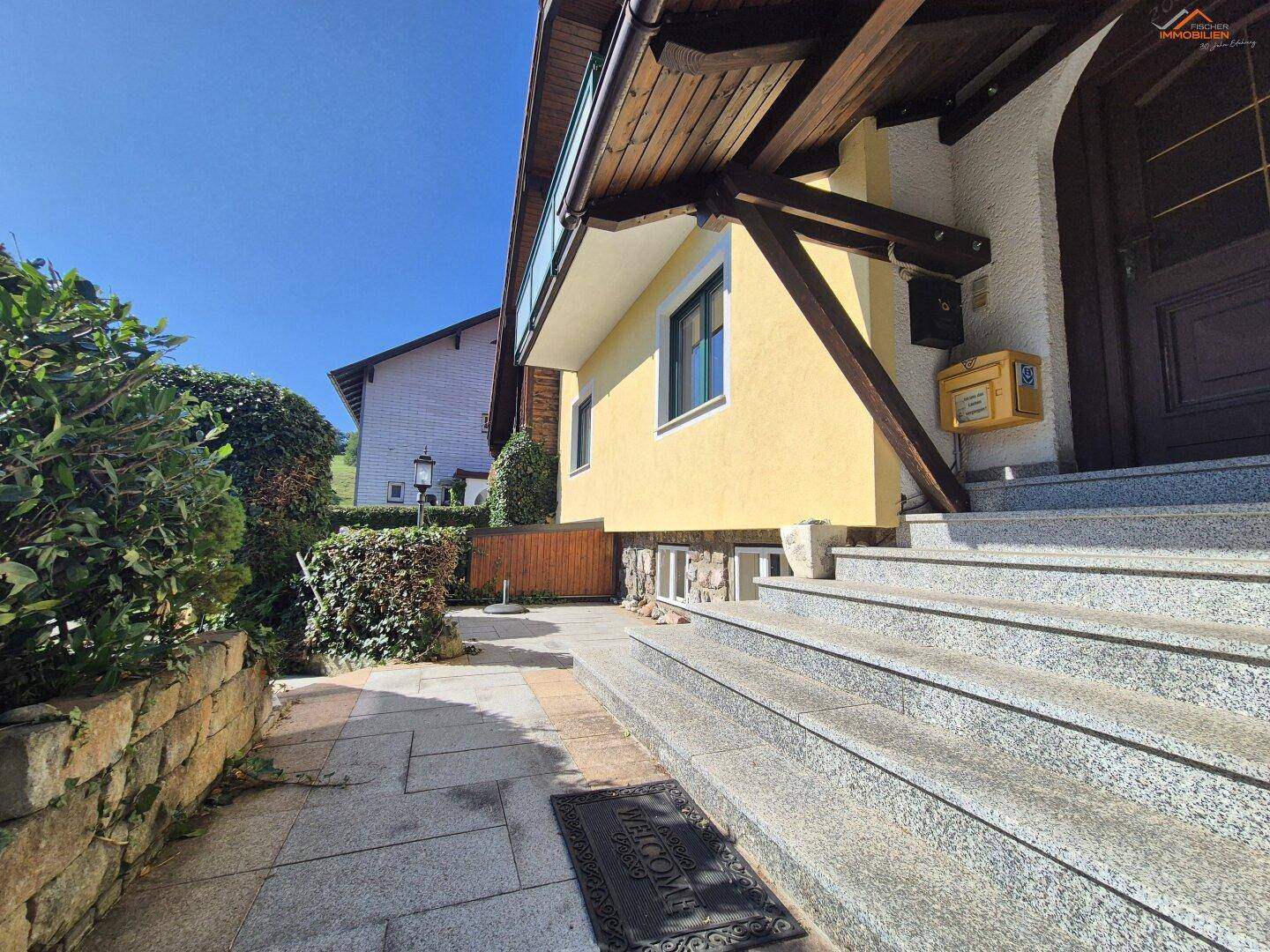 Eingangsbereich des Hauses mit Steintreppe und gepflegter Hecke unter strahlend blauem Himmel.