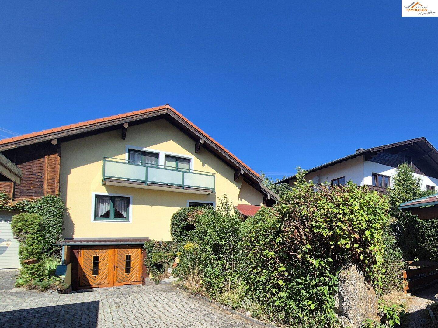 Ansicht des Hauses mit Balkon, gepflasterter Auffahrt und üppiger, grüner Bepflanzung.