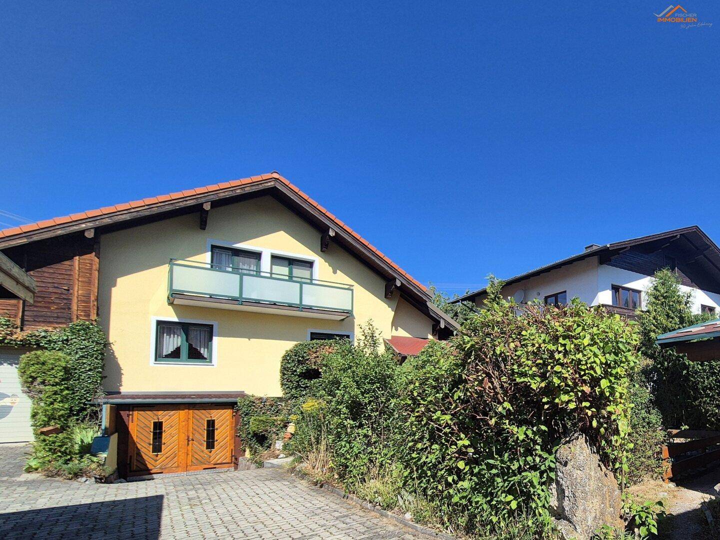 Ansicht des Hauses mit Balkon, gepflegter Hecke und gepflasterter Zufahrt unter blauem Himmel.
