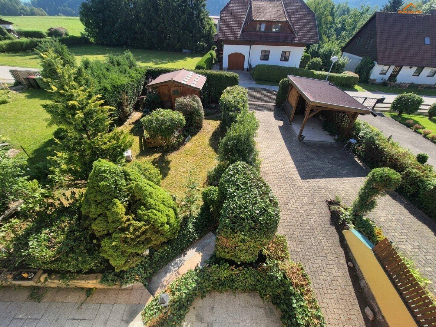 Vogelperspektive des Gartens mit gepflegten Hecken, Bäumen und einem Carport auf der Auffahrt.