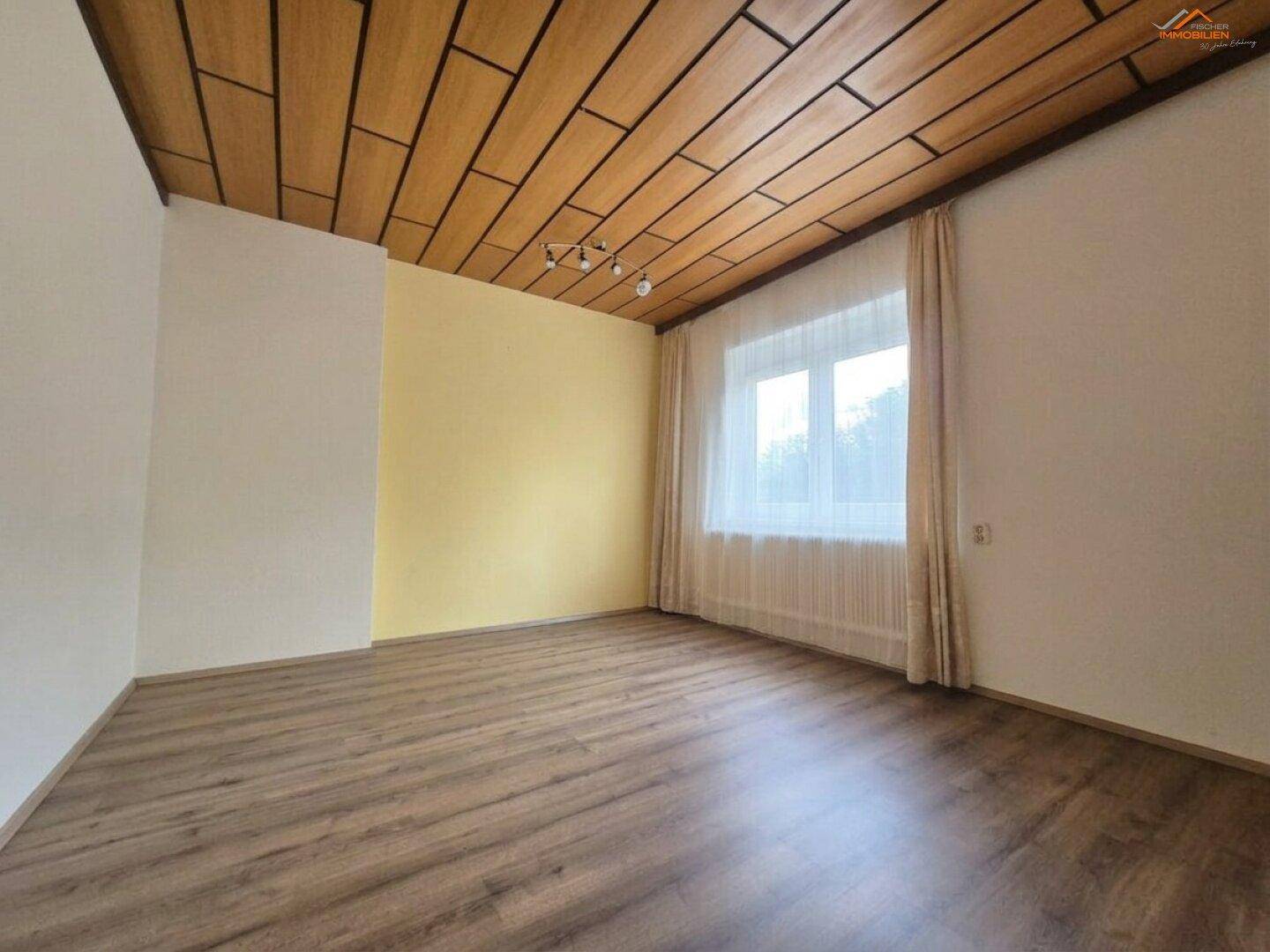Helles Zimmer mit Laminatboden, Holzdecke und großem Fenster, das viel Tageslicht hereinlässt.