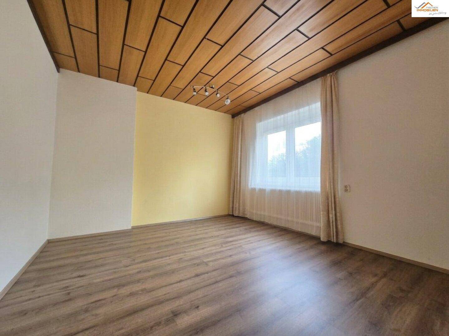 Leeres Zimmer mit Laminatboden, Holzdeckenverkleidung und großem Fenster mit Blick ins Grüne.