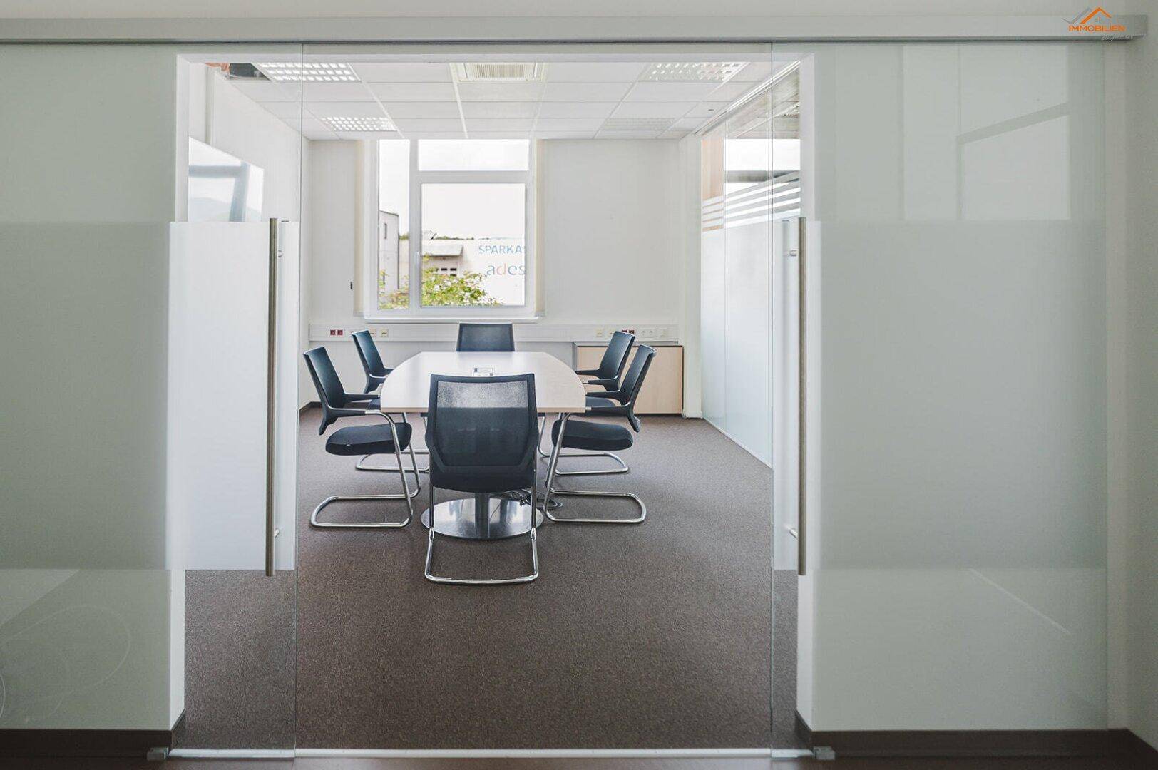 Konferenzraum mit großem Tisch und Stühlen, sichtbar durch moderne Glasschiebetüren, die viel Licht hereinlassen.