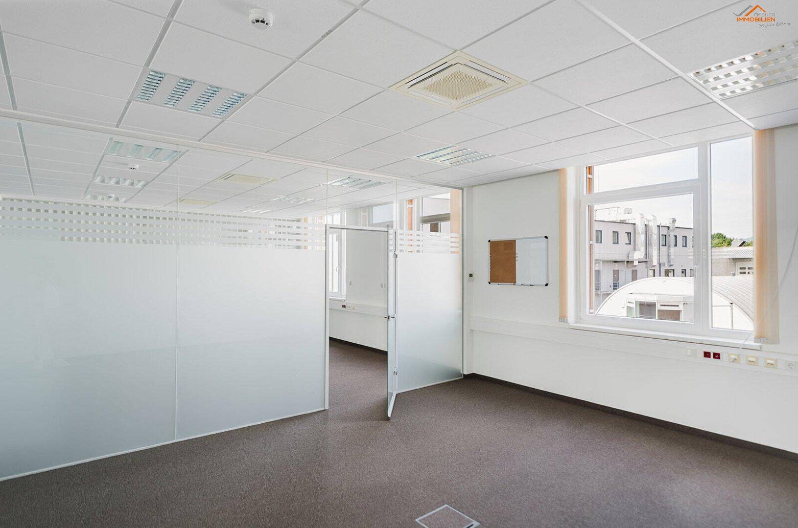 Helles Büro mit Glasabtrennungen, einem Fenster mit Außenblick und einer kleinen Whiteboard-Fläche.