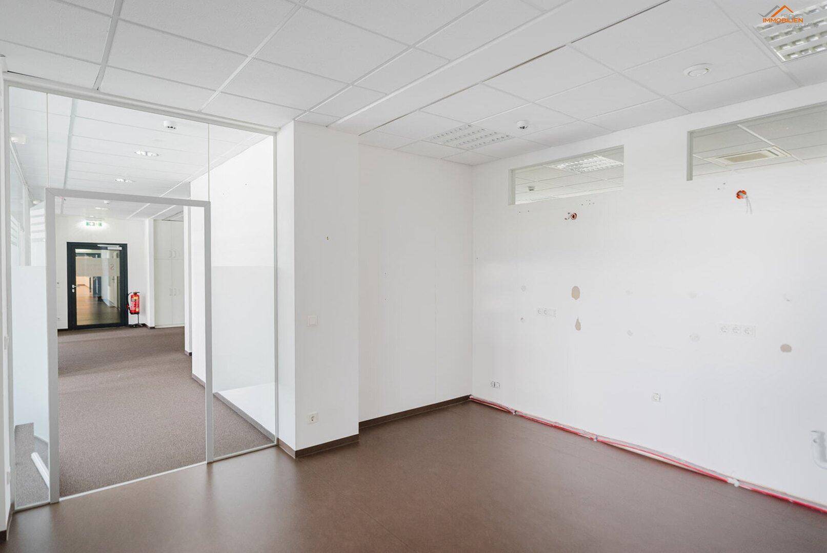 Büroraum mit Glasabtrennung zum Flur, ausgestattet mit braunem Bodenbelag und weißen Wänden.