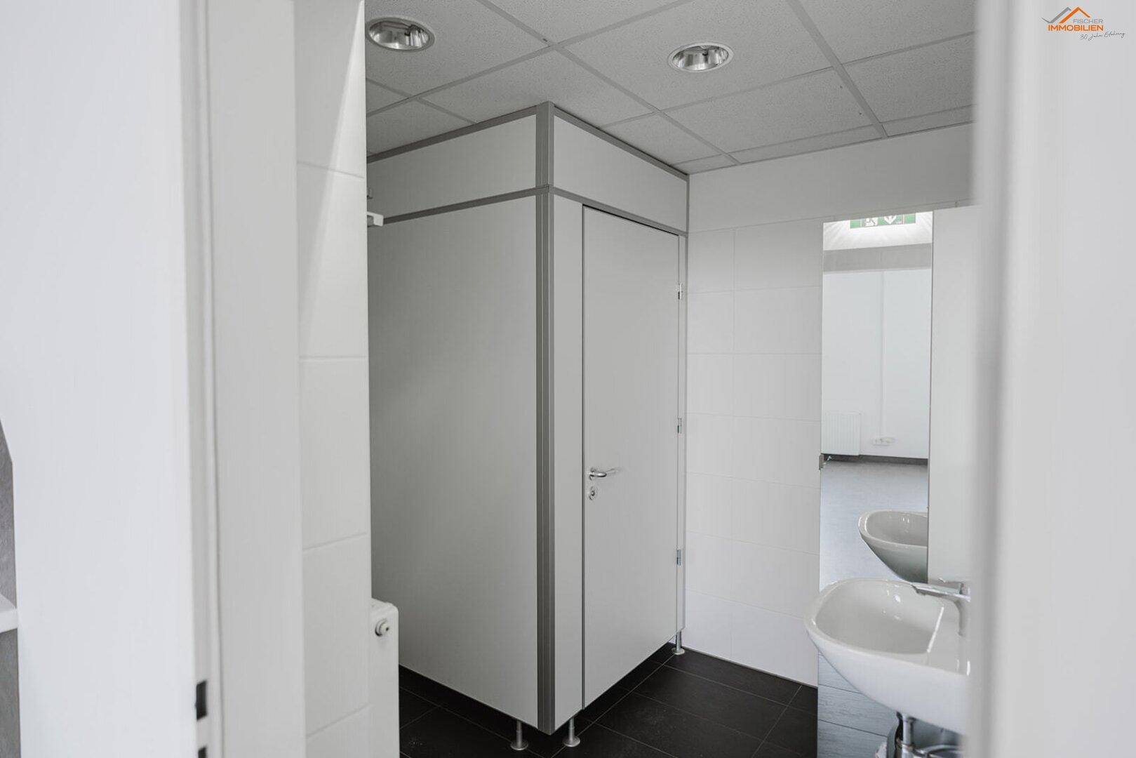 Moderner Sanitärbereich mit weißen Toilettenkabinen und Waschbecken, ausgestattet mit dunklen Fliesen.