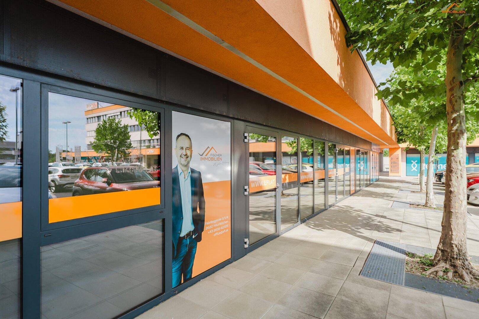 Fassade eines Geschäftsgebäudes mit großen Schaufenstern und einer Werbefläche, umgeben von Bäumen.
