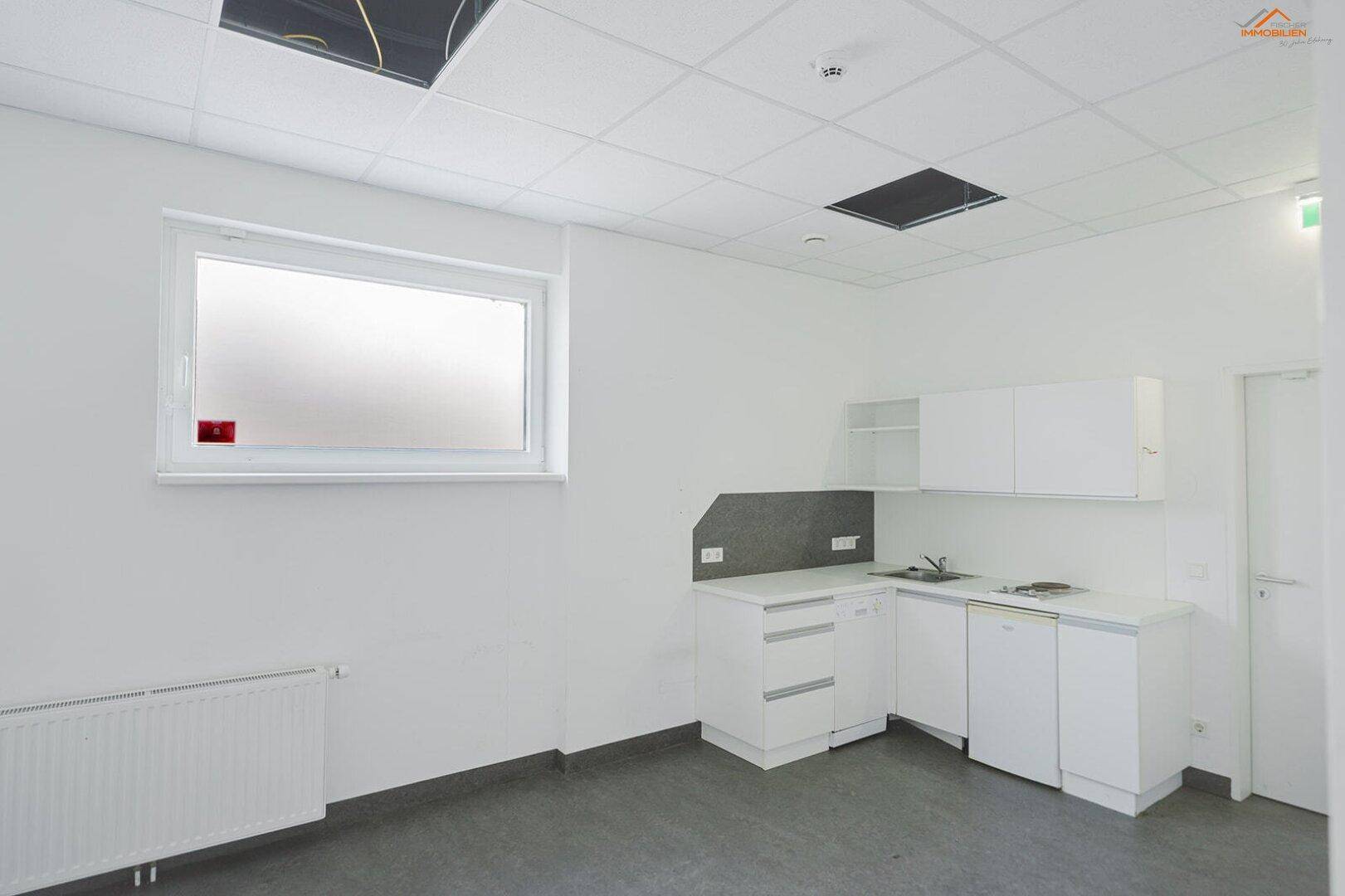 Kleine, funktionale Küchenzeile mit weißen Schränken, Spüle und Fenster in einem Gewerberaum.