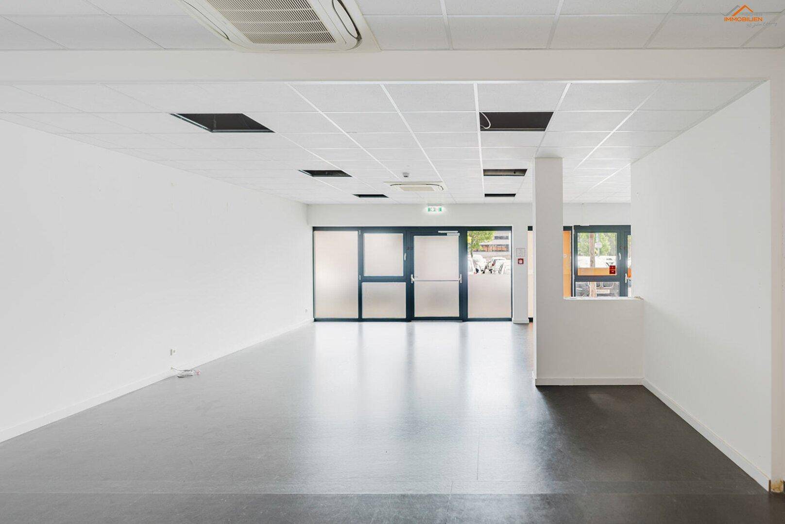 Heller, leerer Gewerberaum mit dunklem Vinylboden, Rasterdecke und großen Fenstern im Hintergrund.