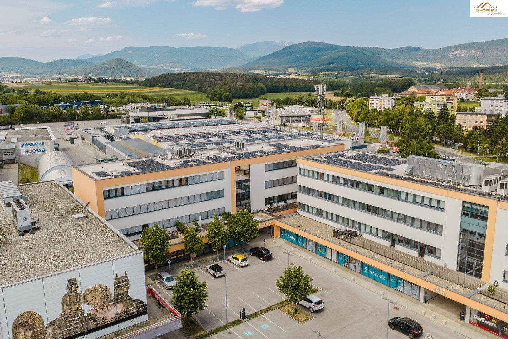 Luftaufnahme eines modernen Geschäftskomplexes mit Solaranlagen auf den Dächern und umliegender Landschaft.