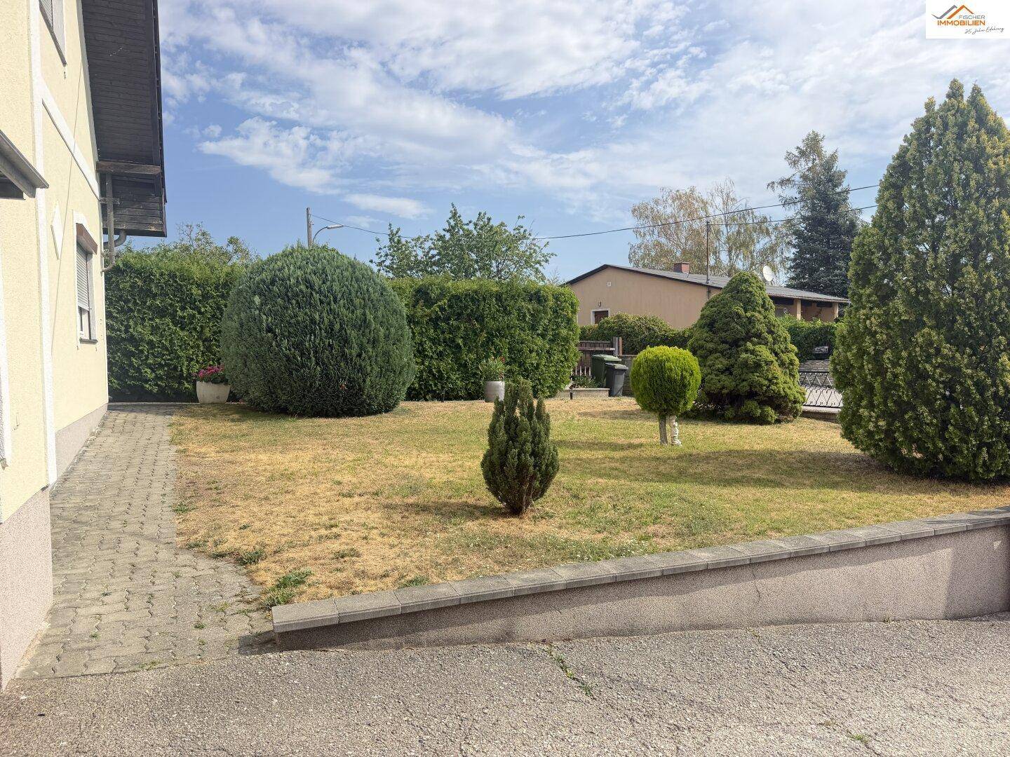 Seitlicher Gartenbereich mit Rasenfläche, Ziersträuchern und gepflastertem Weg.
