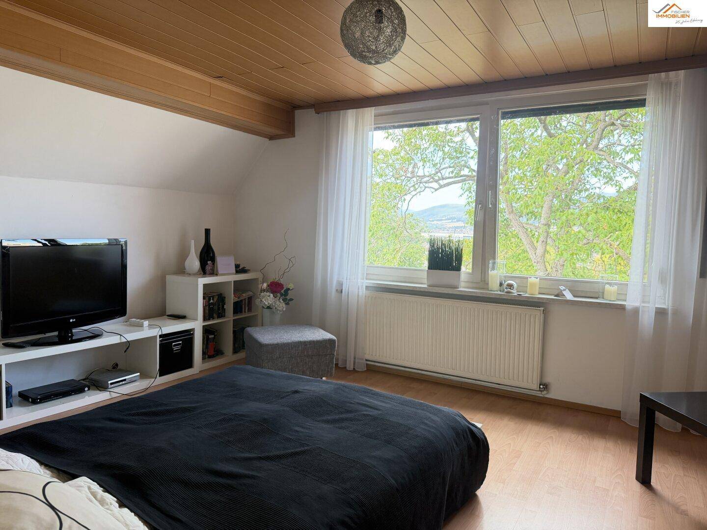 Schlafzimmer im Dachgeschoss mit Holzbalkendecke, großem Fenster und grüner Aussicht.