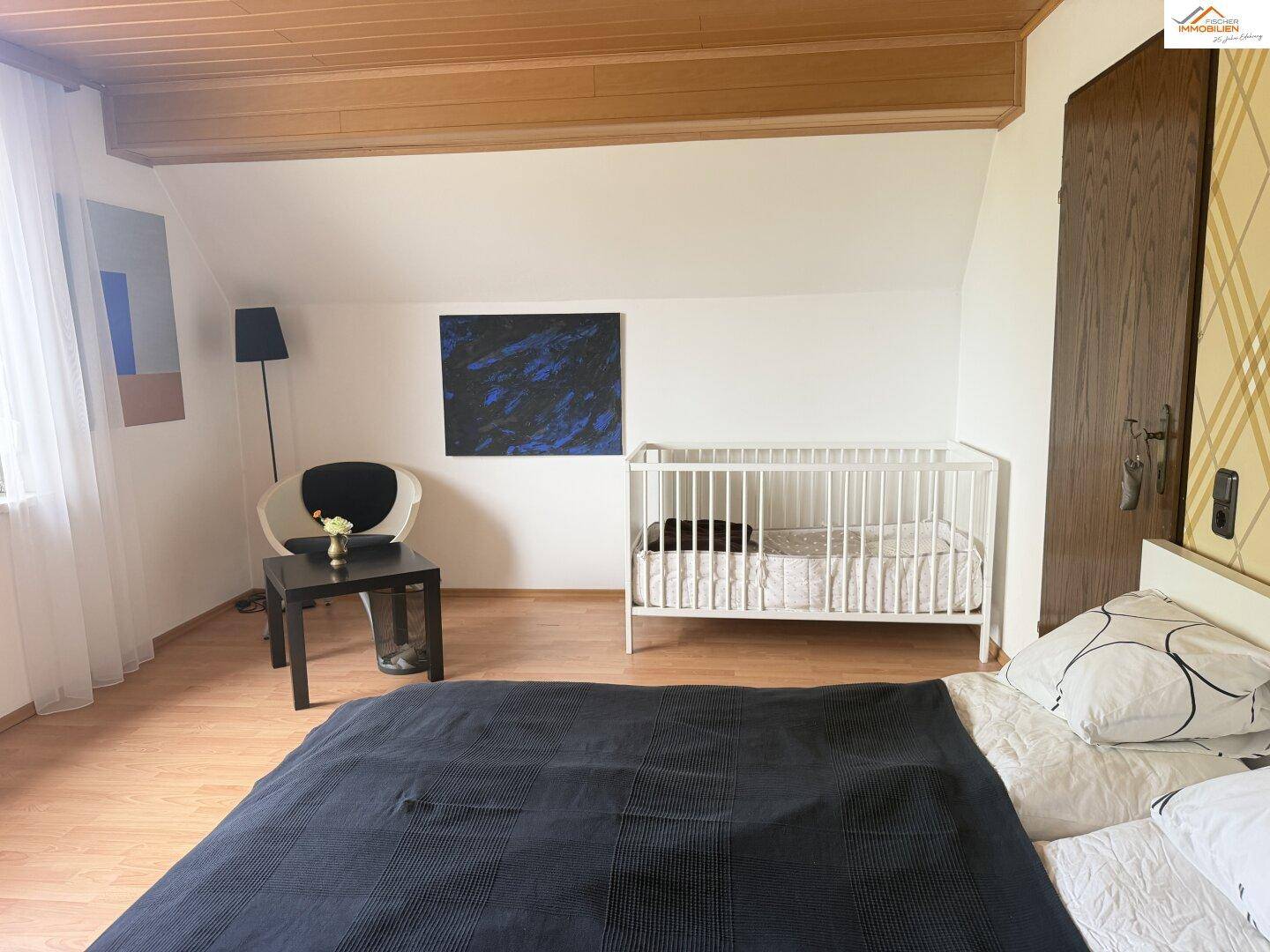 Helles Schlafzimmer mit Babybett, Holzbalkendecke und modernem Kunstwerk.