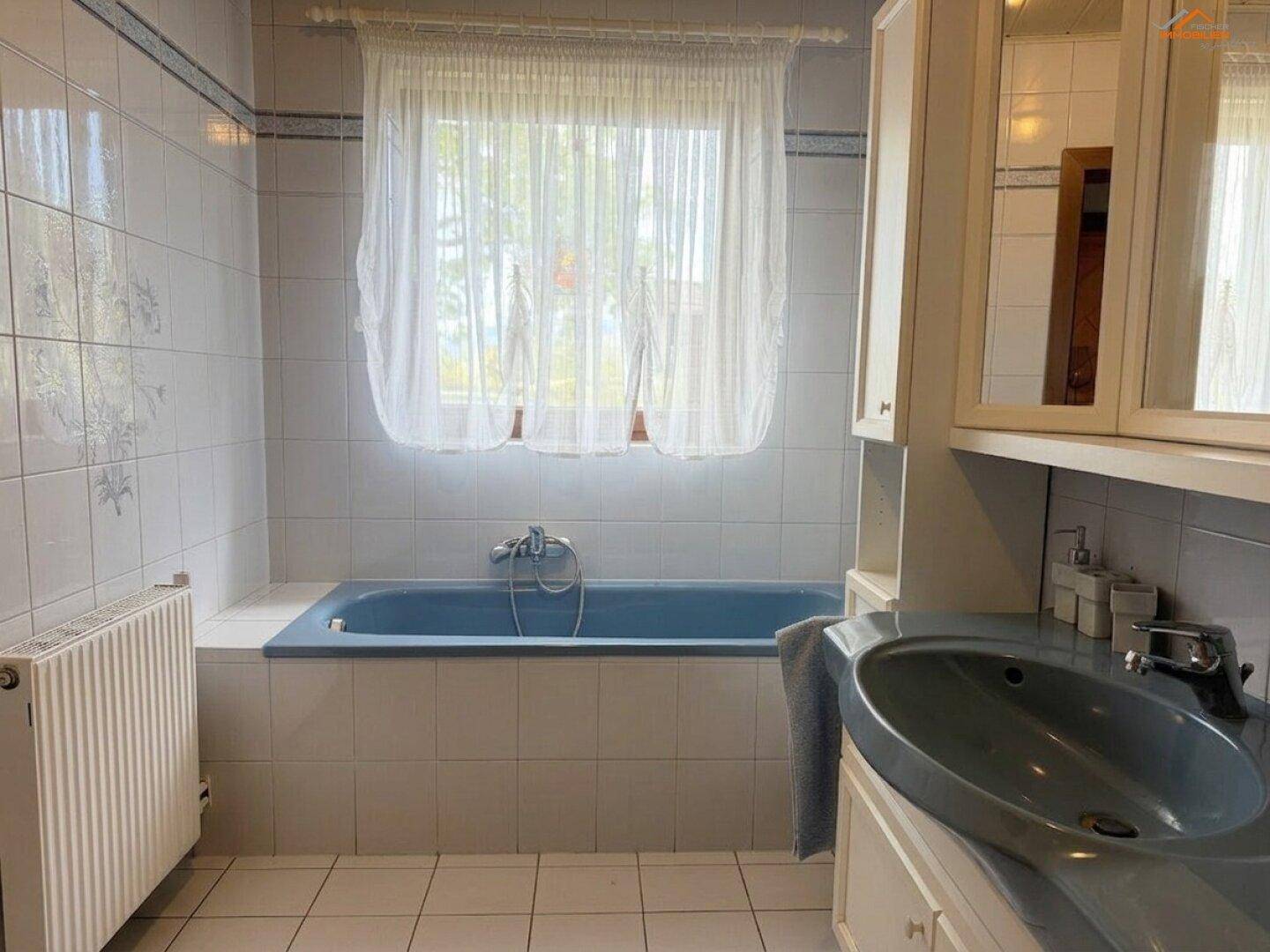 Badezimmer mit Badewanne unter dem Fenster und Heizkörper, alles in hellen Fliesen gehalten.