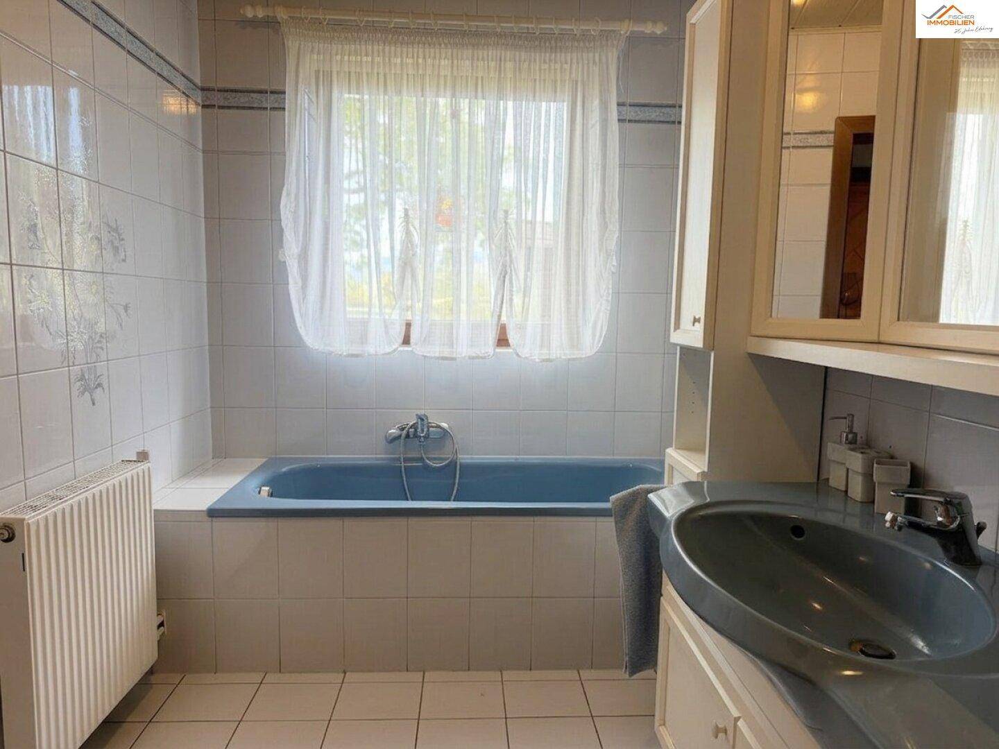 Helles Badezimmer mit blauer Badewanne, Fenster und Heizkörper.