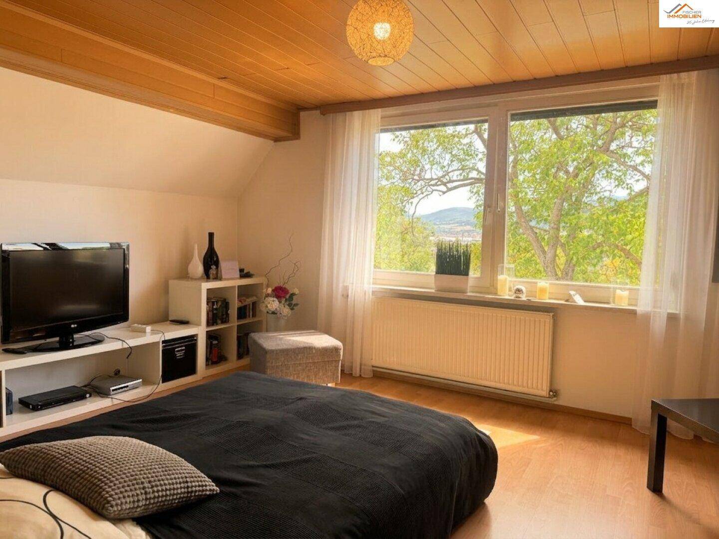 Schlafzimmer mit Holzbalkendecke, großem Fenster und Blick ins Grüne.