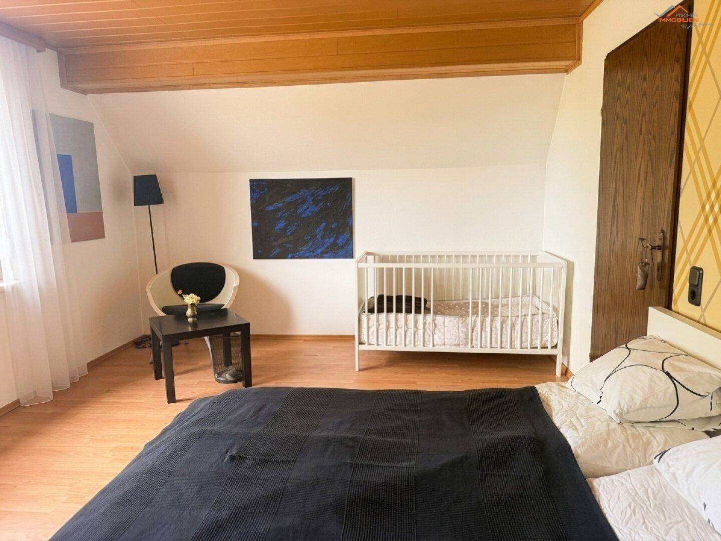 Schlafzimmer mit Babybett, Sitzgelegenheit und Holzbalkendecke, hell und freundlich gestaltet.