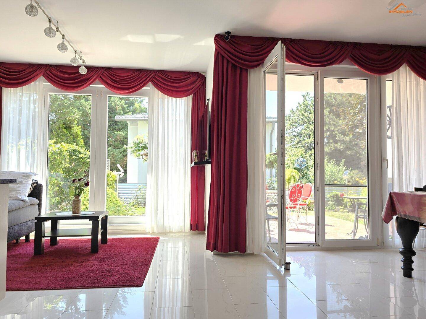 Helles Wohnzimmer mit großen Fenstern, die Zugang zur Terrasse bieten, und eleganten roten Vorhängen.
