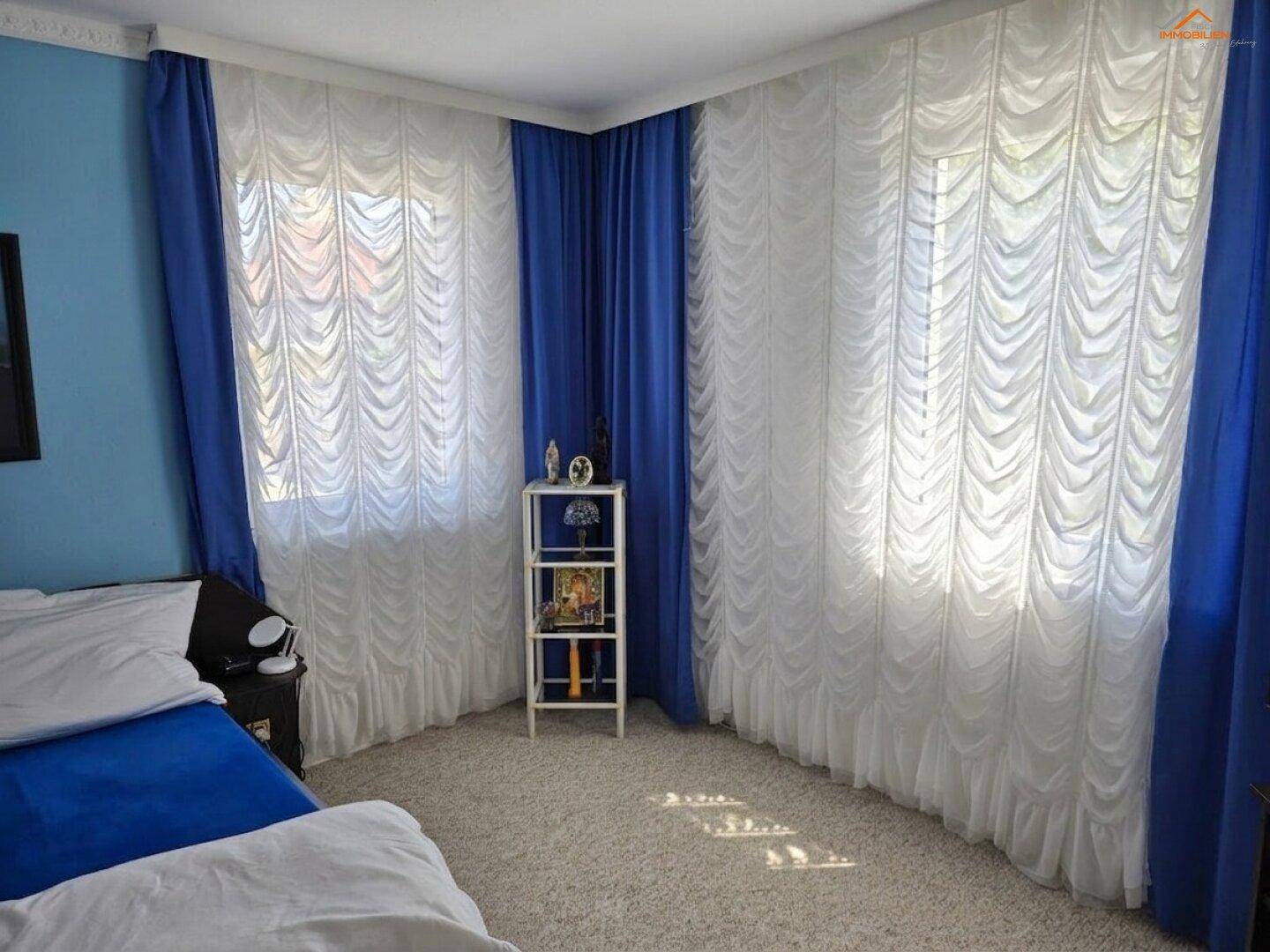 Schlafzimmer mit blauen Vorhängen, Teppichboden und einem Bett, das teilweise sichtbar ist.