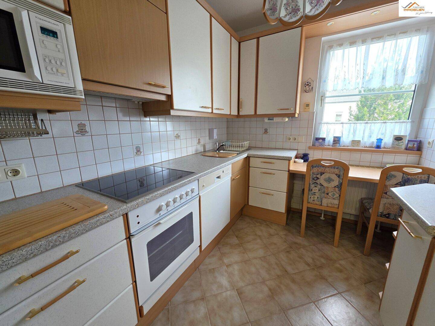 Funktionale Küche mit hellen Holzschränken, Fliesenboden und Essbereich am Fenster.