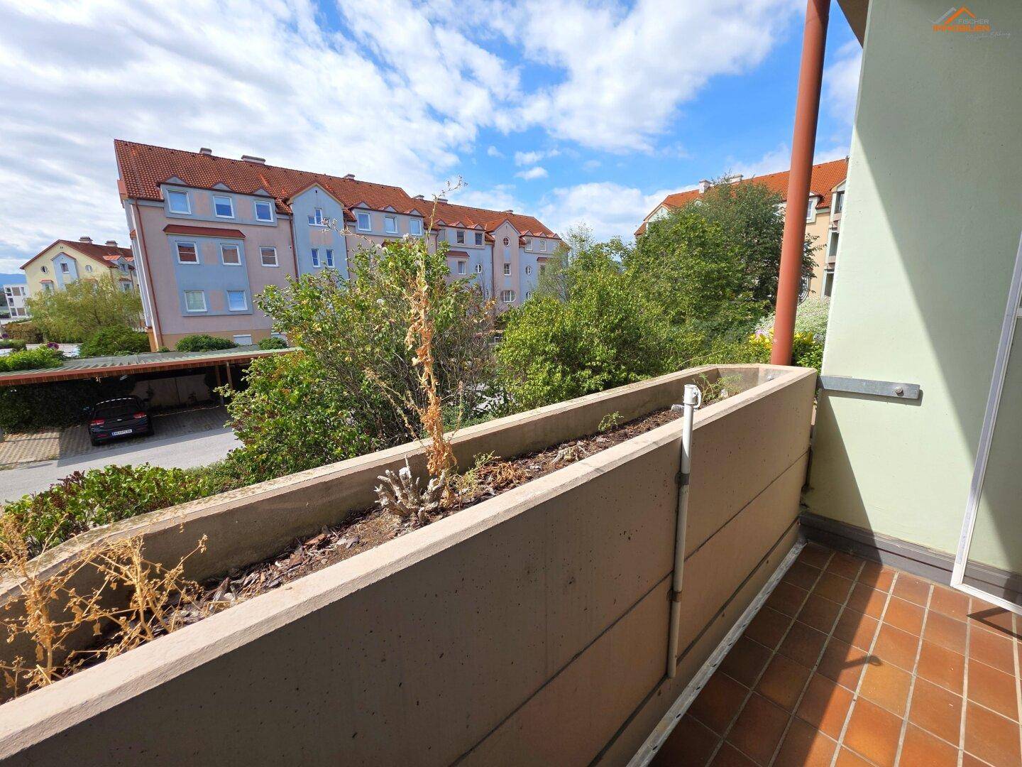 Balkon mit Blick auf die umliegende Wohnbebauung und einen Carport unter blauem Himmel.