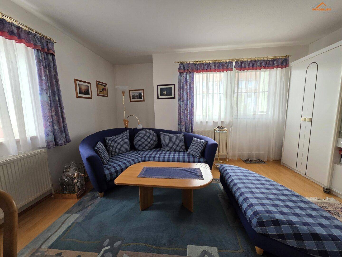 Wohnzimmer mit blauer Eckcouch, Holztisch und großen Fenstern, die viel Licht hereinlassen.