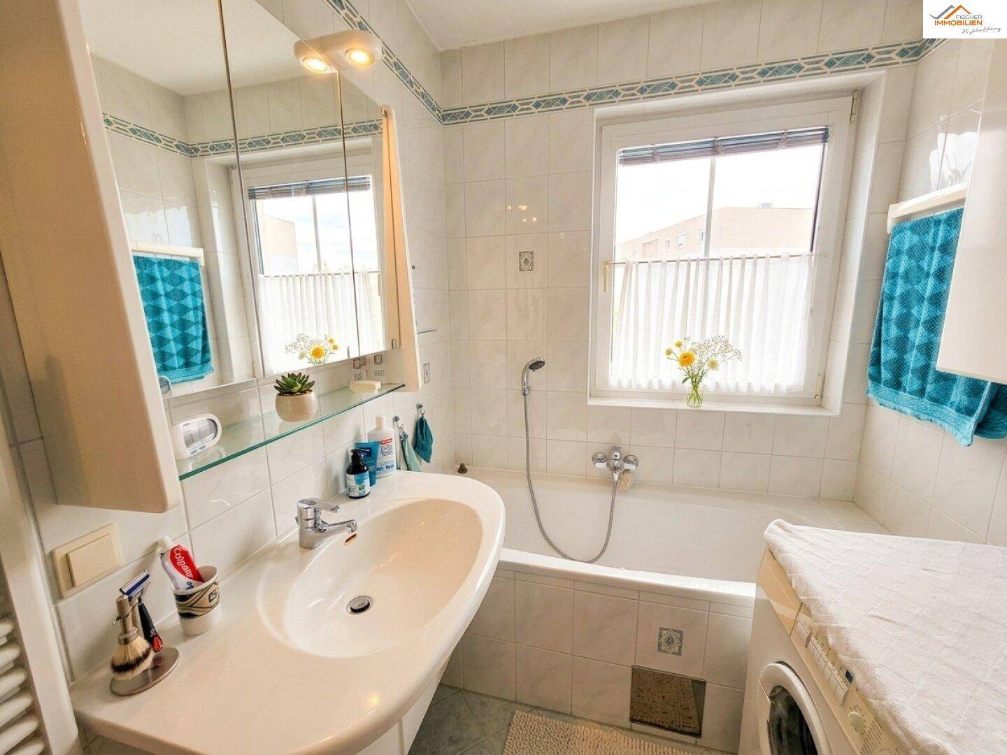 Helles Badezimmer mit Badewanne, Waschbecken und Fenster, ausgestattet mit weißen Fliesen.