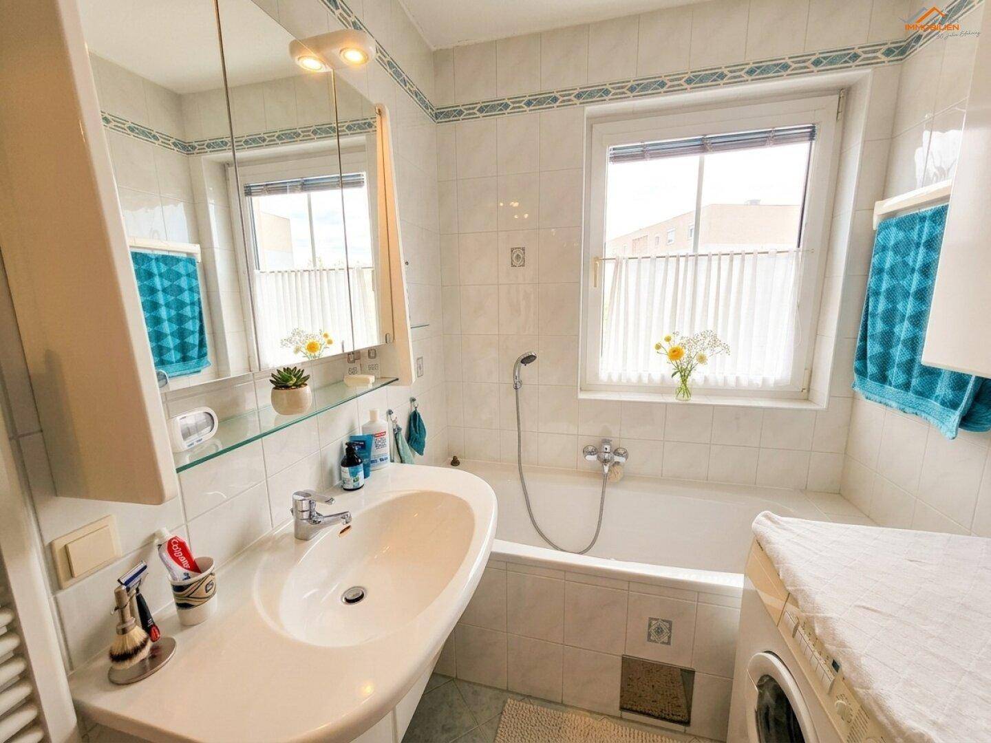 Helles Badezimmer mit Badewanne, Waschbecken und Fenster, ausgestattet mit weißen Fliesen.