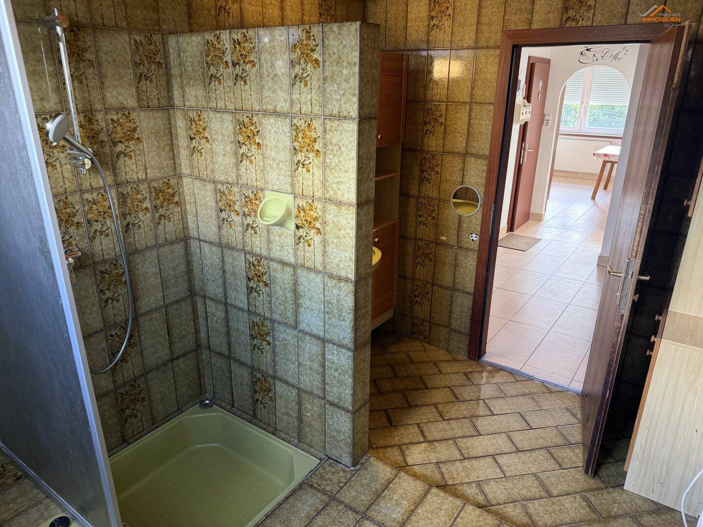 Badezimmer mit Duschbereich, floralen Fliesen und Blick in den angrenzenden Flur, typisch für die 80er Jahre.