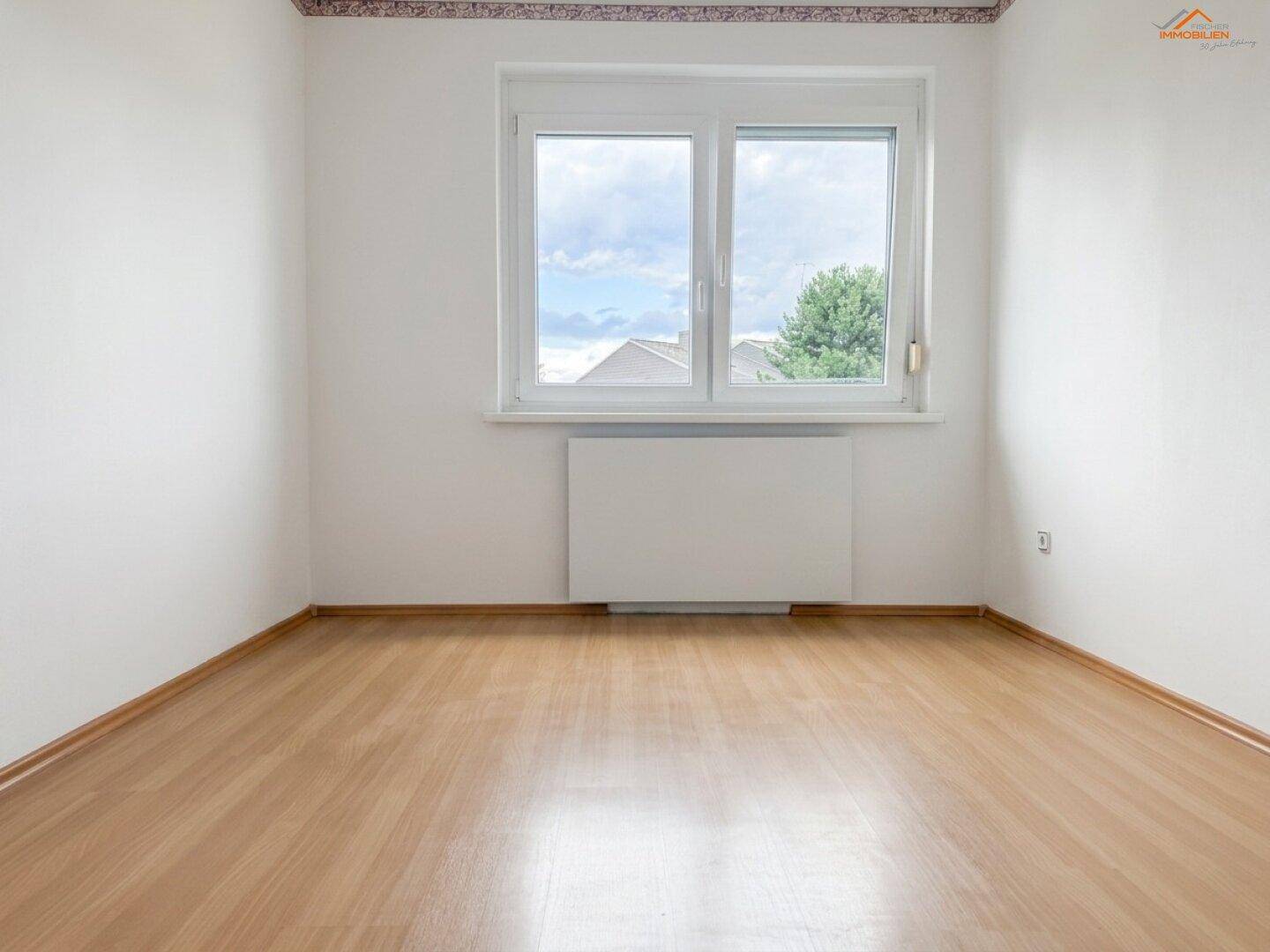 Helles Zimmer mit Laminatboden, weißen Wänden und einem großen Fenster, das viel Tageslicht hereinlässt.