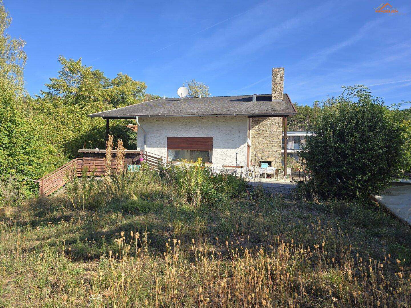 Kleines Haus mit Holzterrasse, Kamin und umgeben von üppiger, naturbelassener Vegetation.