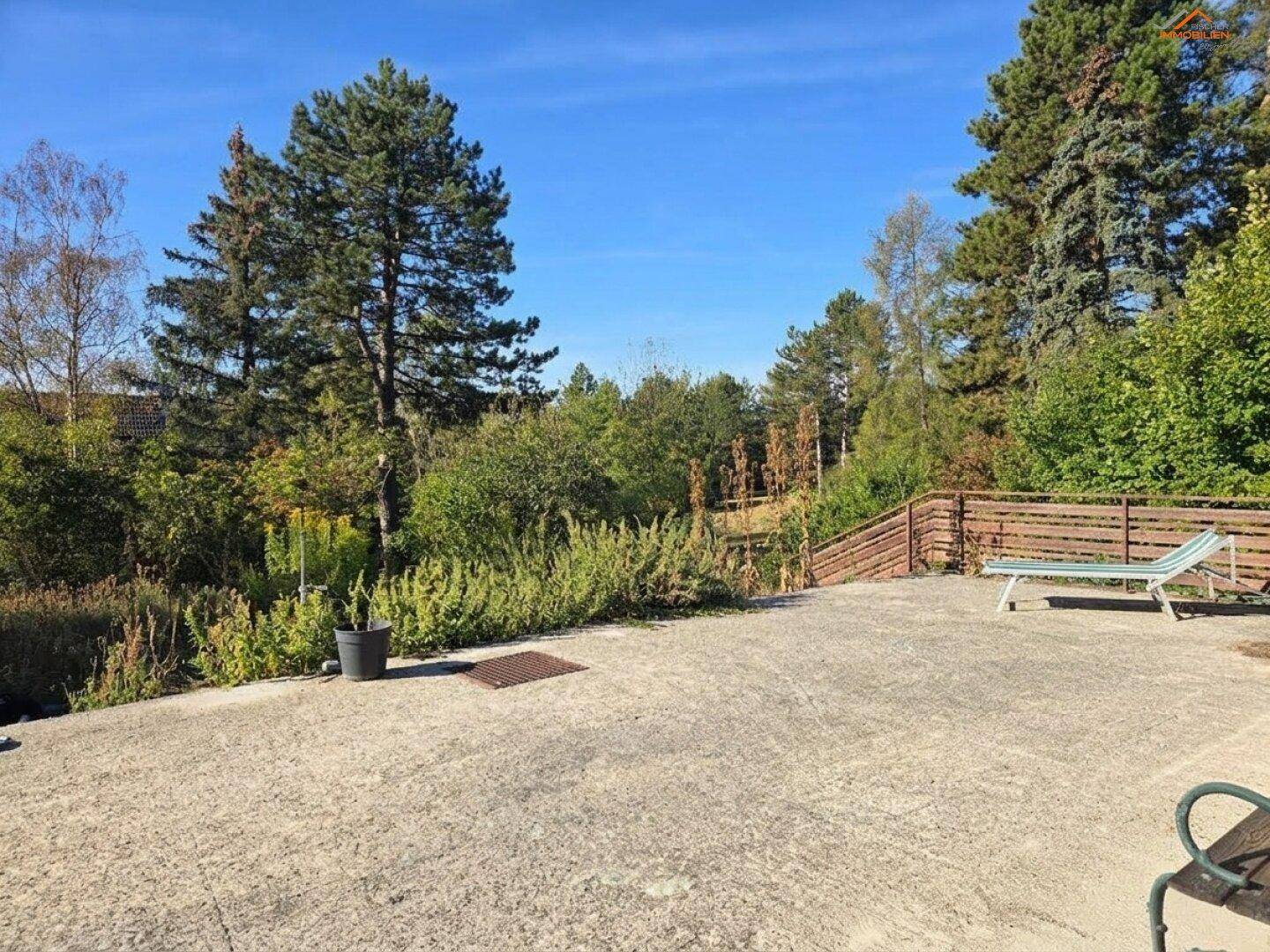 Große gepflasterte Terrasse mit Liegestuhl, umgeben von üppigem Grün und Bäumen.