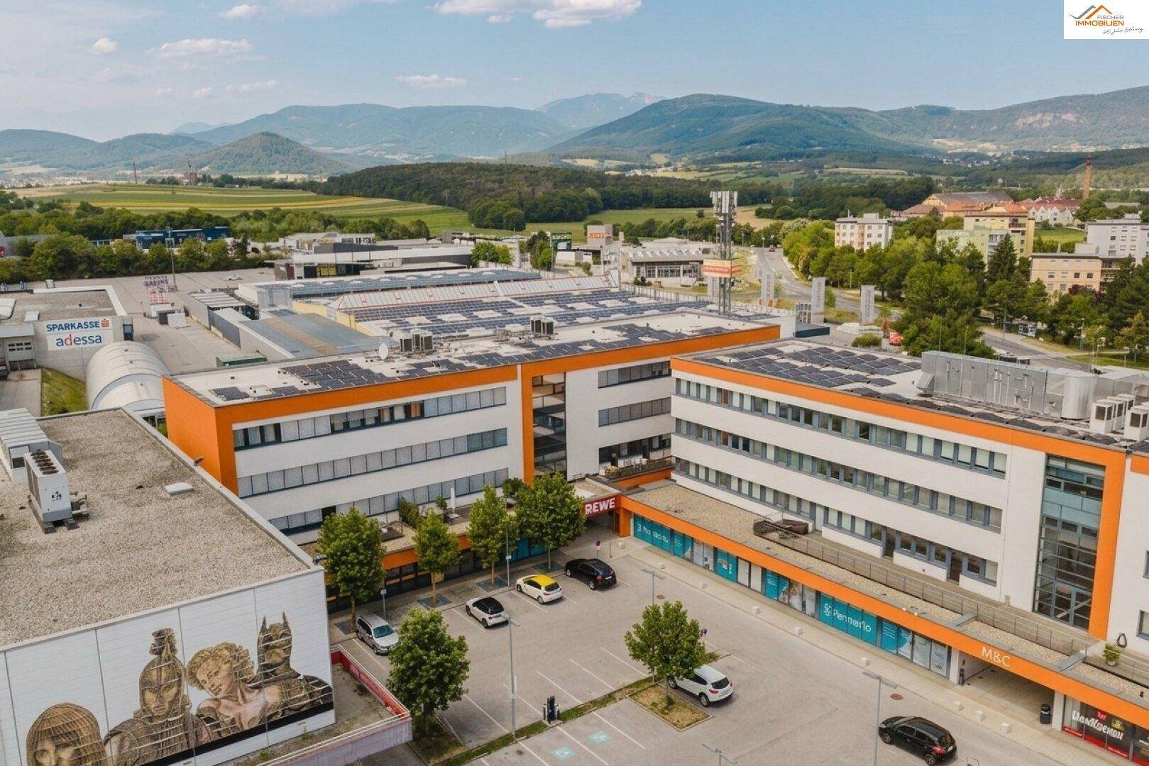 Luftaufnahme eines Gewerbekomplexes mit modernen Gebäuden, orangefarbenen Fassaden und Solaranlagen auf den Dächern. Parkplätze und Geschäfte sind sichtbar, umgeben von grüner Landschaft und Bergen.