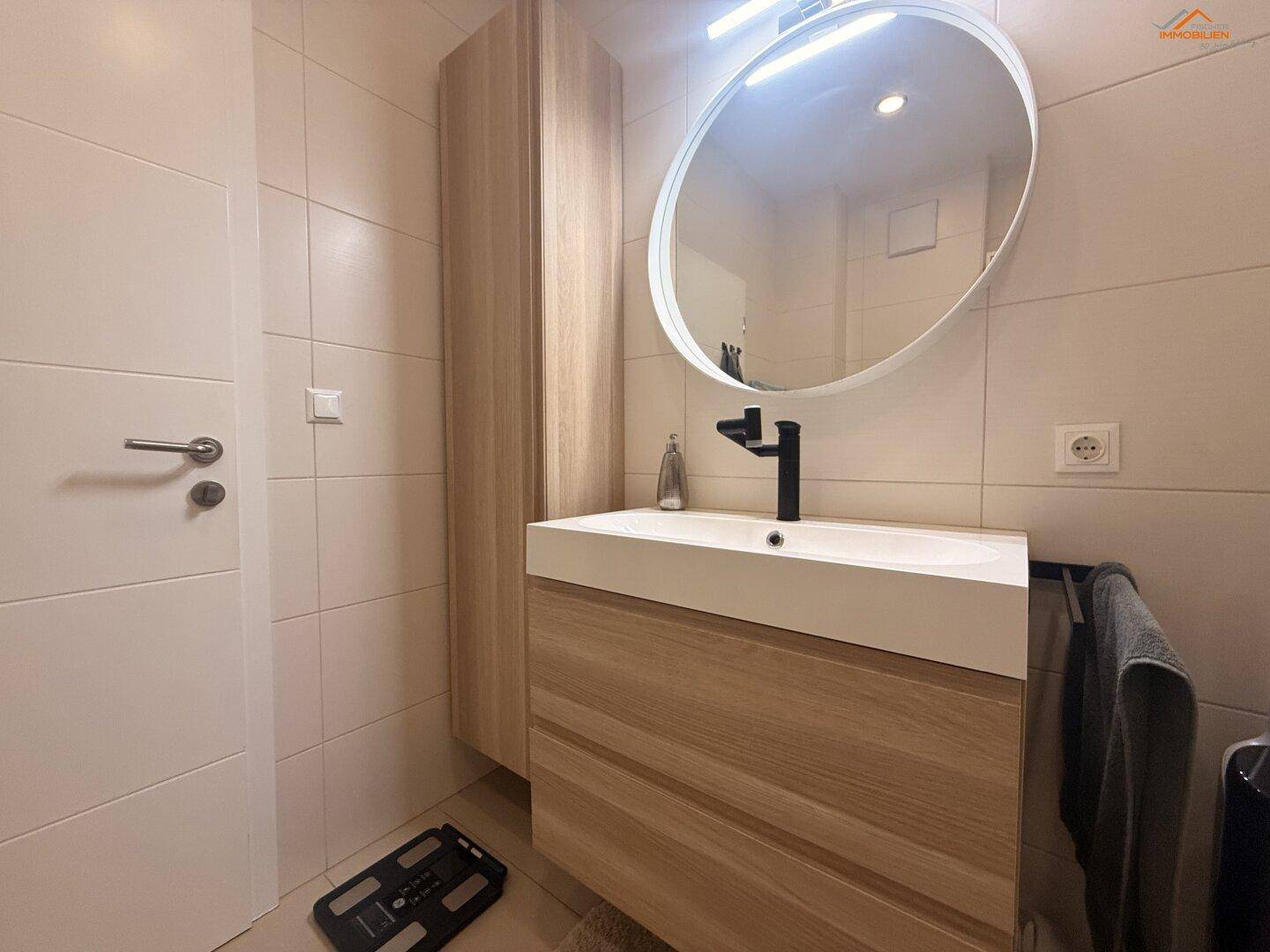 Badezimmer mit modernem Waschtisch, rundem Spiegel und hellen Fliesen.