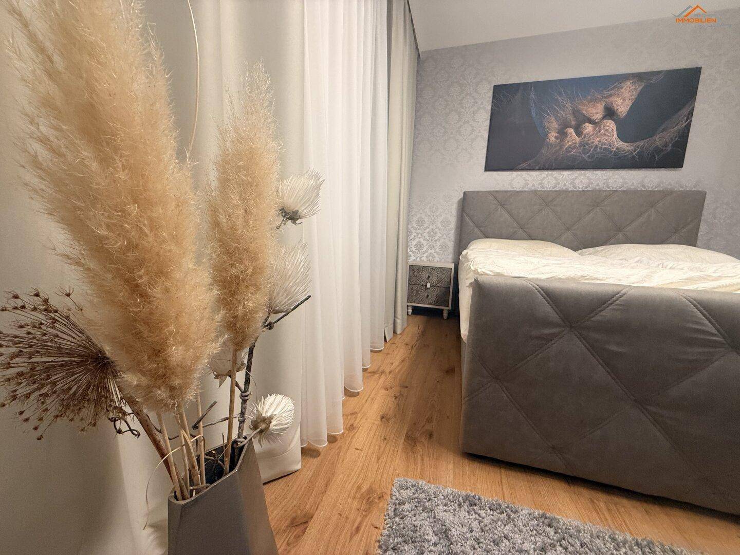Schlafzimmer mit gepolstertem Bett, Holzboden und dekorativen Trockenblumen.