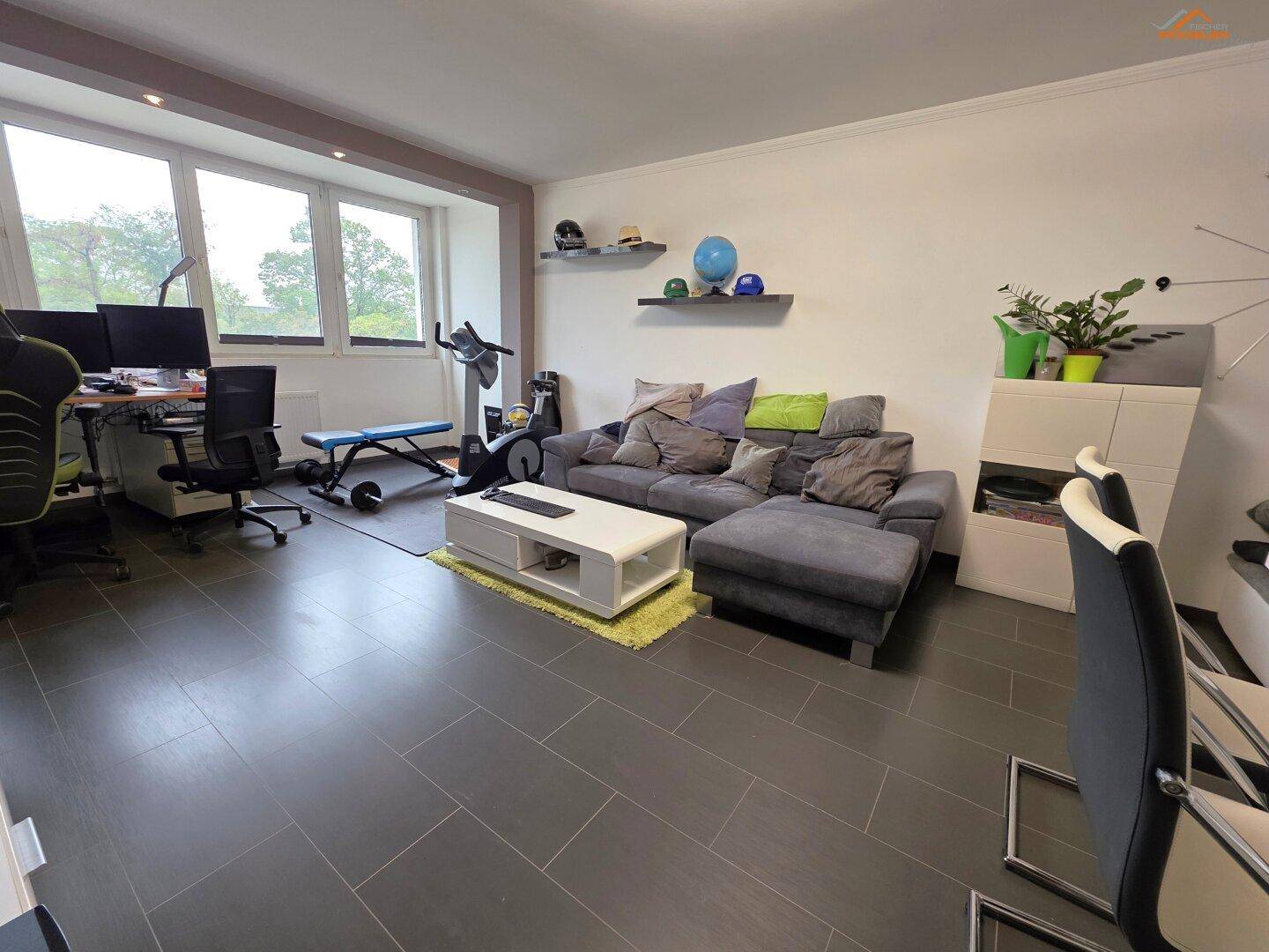 Geräumiges Wohnzimmer mit dunklen Fliesen, Sofa, Schreibtisch und Fitnessgeräten.