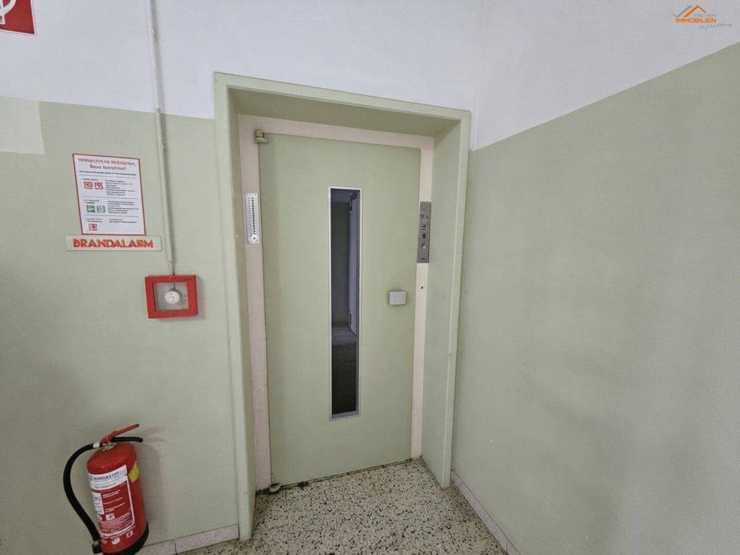 Flur im Gebäude mit Aufzug, Feuerlöscher und Hinweisschildern.