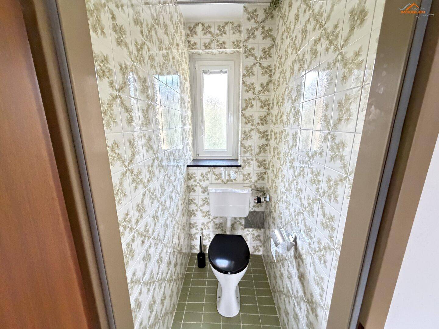 Separate Toilette mit gemusterten Fliesen, einem Fenster und grünem Fliesenboden.