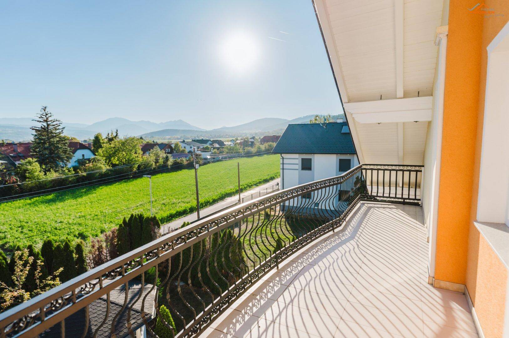 Sonniger Balkon mit schmiedeeisernem Geländer und weitem Blick auf die grüne Landschaft und Berge.
