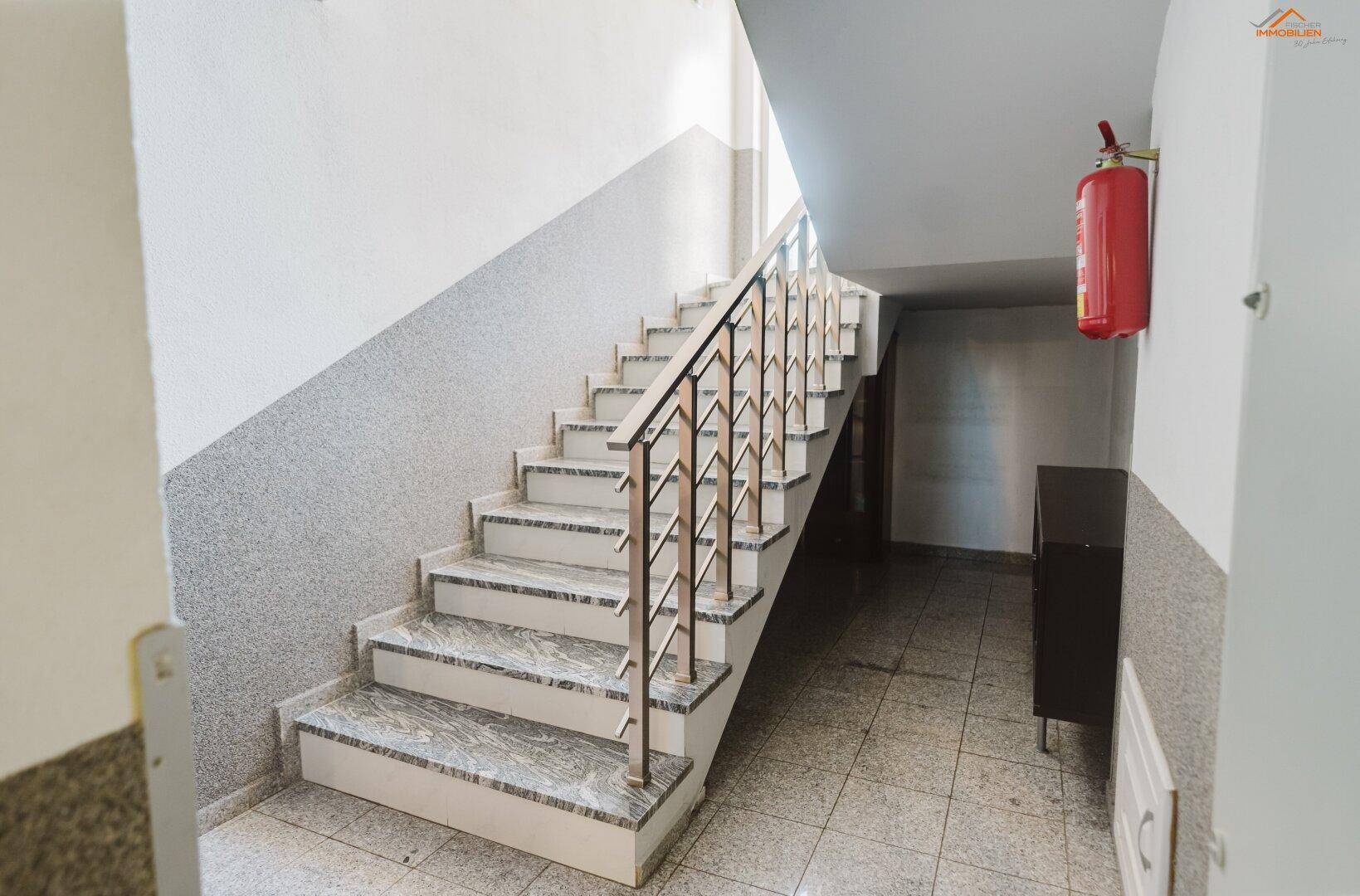 Treppenhaus mit Marmorstufen, Edelstahlgeländer und einem Feuerlöscher an der Wand.