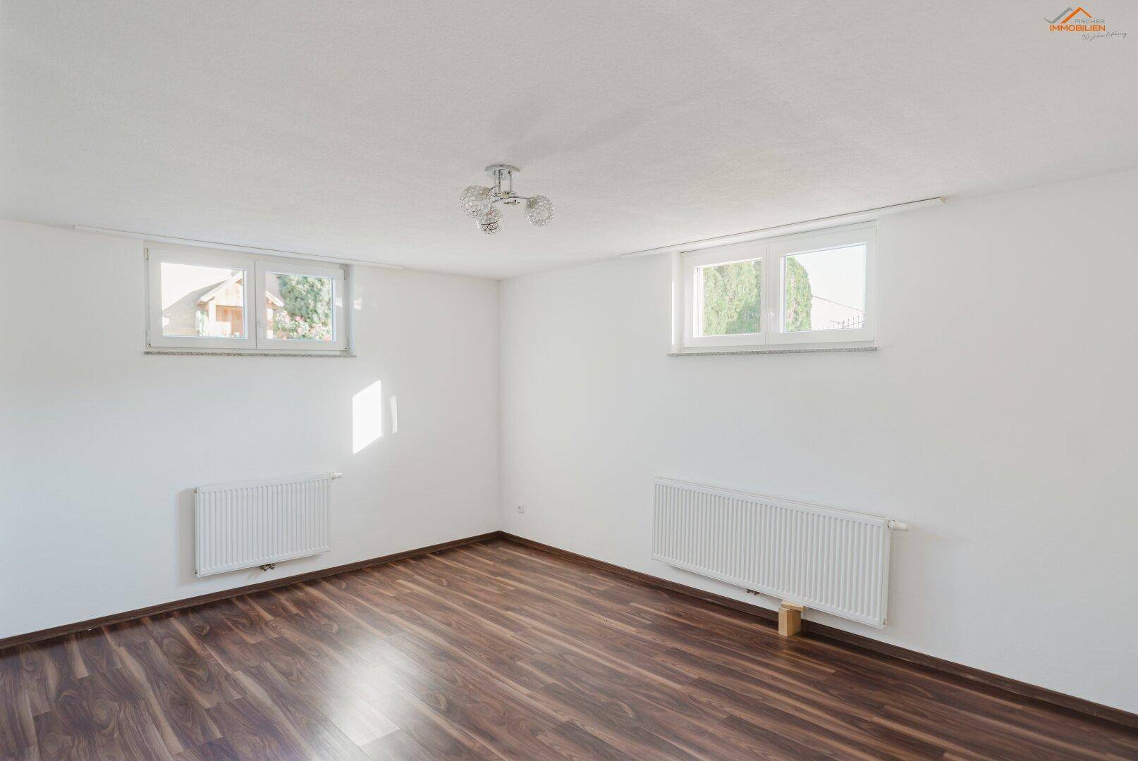 Helles Schlafzimmer mit dunklem Holzboden und zwei Fenstern, die viel Tageslicht hereinlassen.