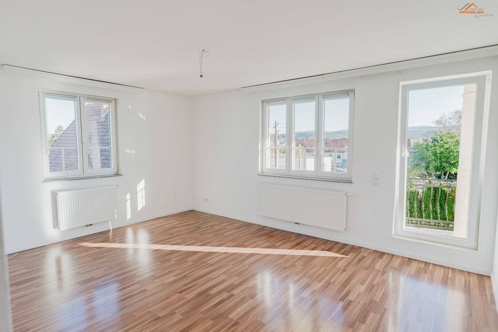 Heller Wohnraum mit Holzboden, großen Fenstern und direktem Zugang zum Balkon.