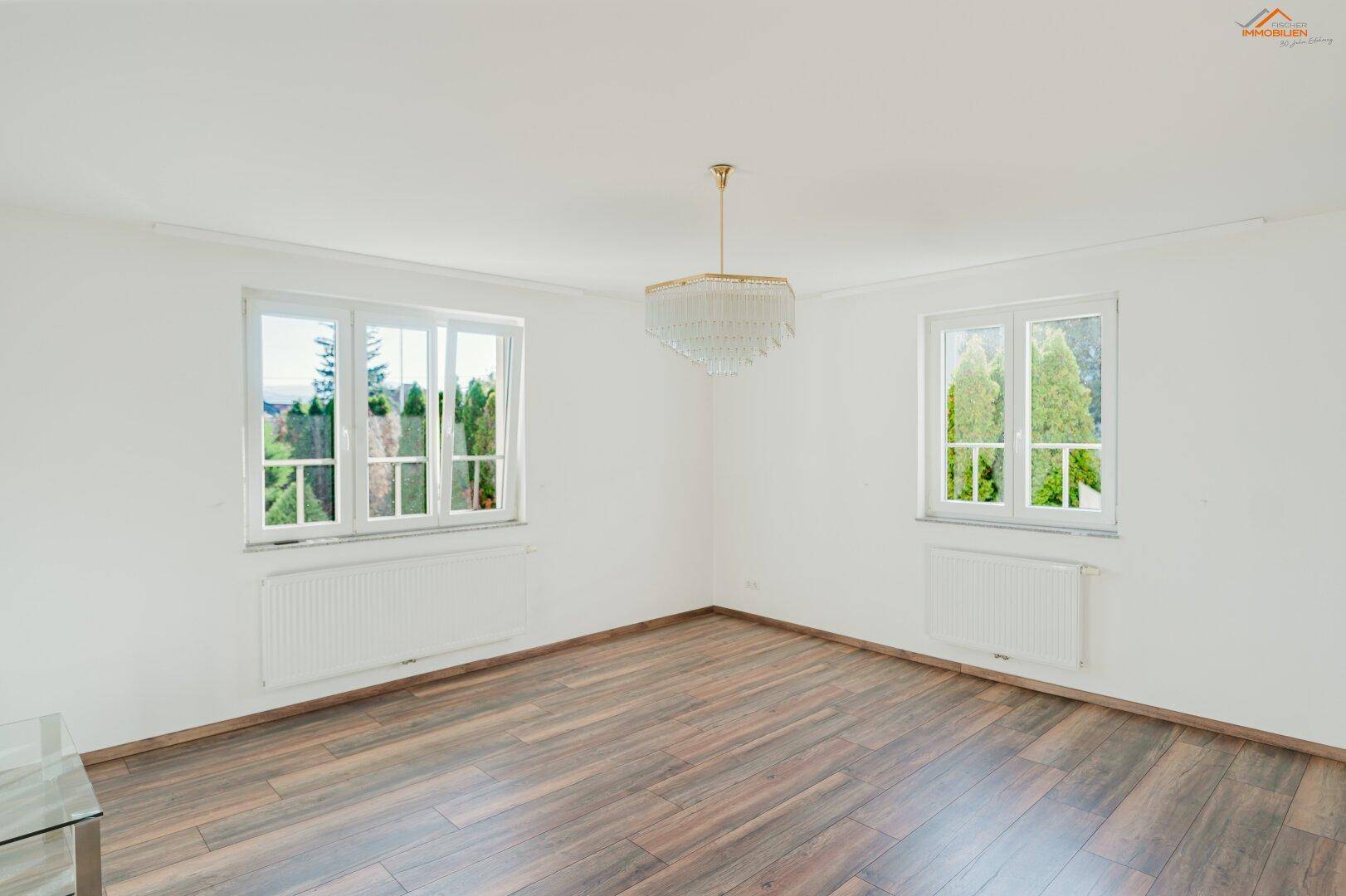 Geräumiger Raum mit dunklem Holzboden, zwei großen Fenstern und einem eleganten Kronleuchter.