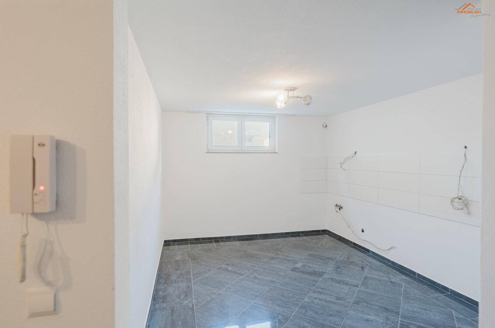 Leerer Küchenraum mit Fliesenboden, einem Fenster und vorbereiteten Anschlüssen für eine neue Küche.